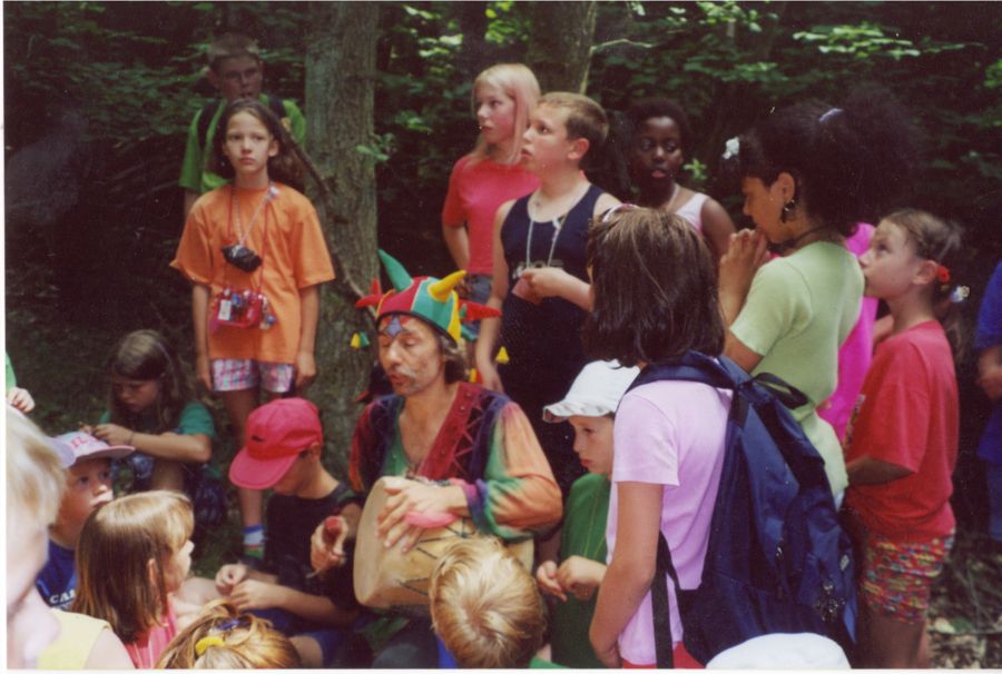 Micha mit Narrenkappe und Trommel im Wald, umringt von lauschenden Kindern