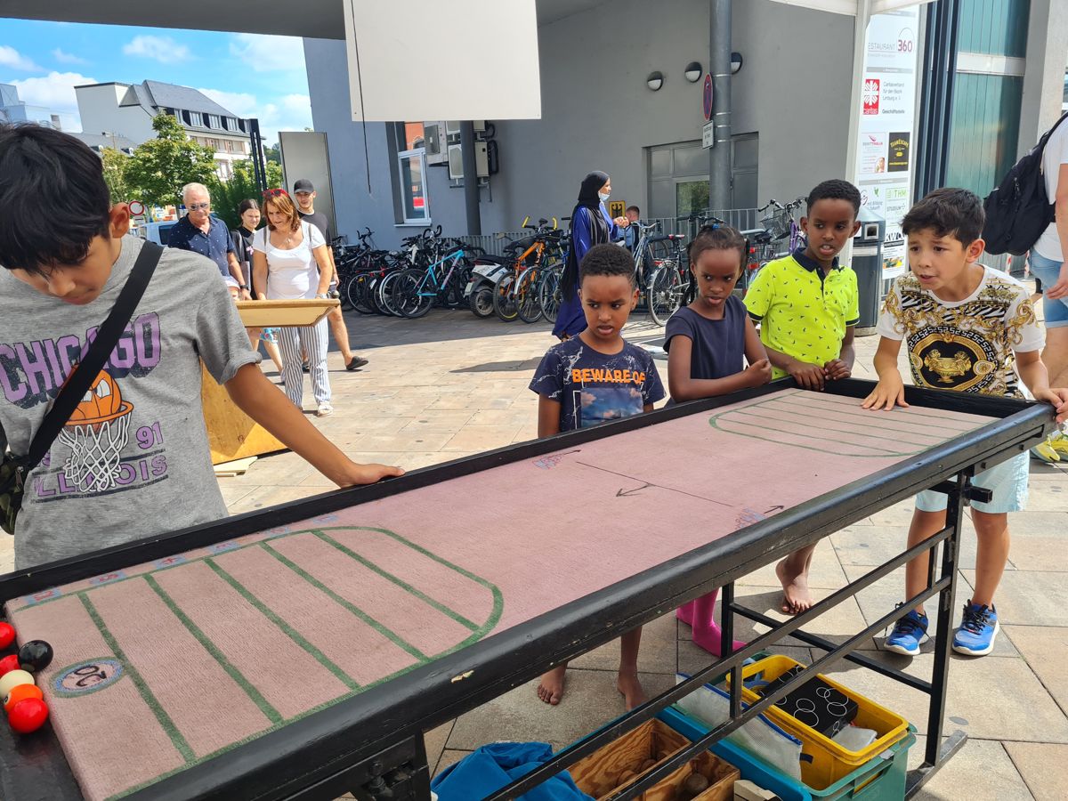 Kinder stehen gespannt am langen Coolball-Tisch und verfolgen, wie ein Junge seine Kugel spielt