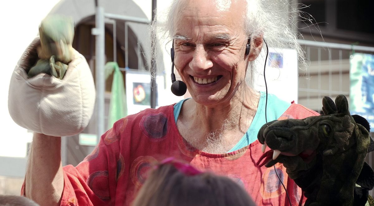 Micha lacht mit Headset, in der einen Hand ein Drachenei im Stoffbeutel, in der anderen eine Drachenpuppe