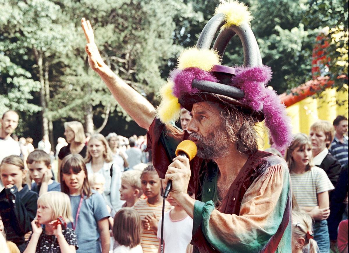 Micha mit Mikrofon und buntem Fantasiehut moderiert vor vielen Kindern